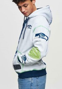 Vit hoodie med blå och gröna detaljer, med en Seahawks-logotyp och text på ärmen. Huva design med ribbad nederkant.