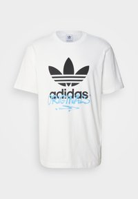 T-shirt bianco in cotone con un grande logo Adidas nero con tre strisce e testo in stile graffiti blu che dice "ORIGINALS" sotto.