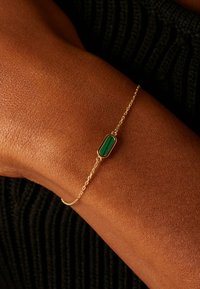 Gouden kettingarmband met een ovale groene steen als middelpunt. De ketting is delicaat met een zachte textuur tegen de huid.