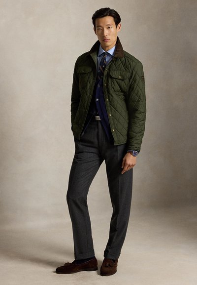 Hombre con chaqueta acolchada verde, camisa azul con corbata, pantalones grises y mocasines marrones, de pie frente a un fondo liso.