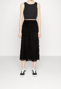 Černý zkrácený top bez rukávů v kombinaci s černou sukní ke kolenům, která má strukturovaný vzor. Doplňují to bílé a námořnické tenisky.