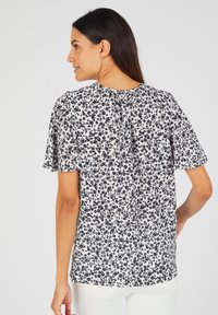 Korte mouwen blouse in een witte stof met een zwart bloemenpatroon. Heeft een ronde halslijn en een rugplooitje voor extra detail.