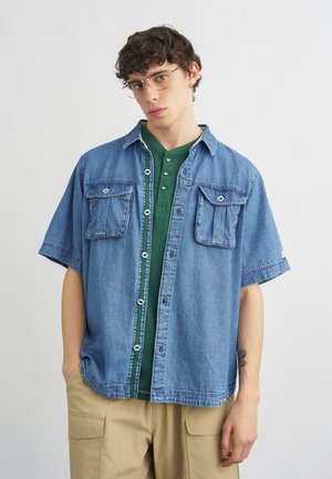 Mladý muž s kučeravými vlasmi nosí priehľadné okuliare, zelené henley tričko, otvorenú krátku modrú rifľovú bundu a béžové cargo nohavice.