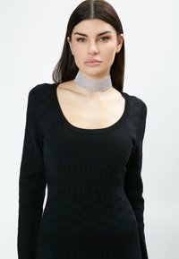 Schwarzer, langärmliger Pullover mit strukturierten Mustern, die "INFLUENCER" buchstabieren, und einem runden Ausschnitt, kombiniert mit einem funkelnden silbernen Choker-Halsband.