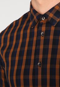 Chemise boutonnée en motif à carreaux bleu marine et orange. Comprend un col standard et des boutons foncés sur la patte de boutonnage frontale.