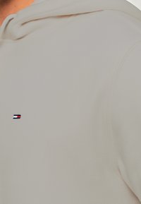 Tissu beige avec un petit logo rectangulaire comprenant des bandes horizontales blanches, rouges et bleu foncé sur la zone de la poitrine.