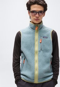 Νέος άνδρας που φοράει γυαλιά ηλίου, γαλάζιο φλις αμάνικο με πορτοκαλί φερμουάρ και μαύρο μακρυμάνικο πουκάμισο, στέκεται με τα χέρια στις τσέπες.