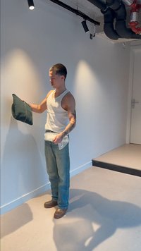 Mężczyzna w koszulce bez rękawów trzyma czarną saszetkę i przezroczystą plastikową torbę, stojąc w minimalistycznym pokoju z białymi ścianami i odsłoniętymi rurami na suficie.