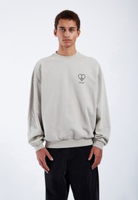 Felpa color beige chiaro con polsini e orlo a costine. Sul davanti presenta un piccolo grafico scuro a forma di cuore con del testo sottostante.