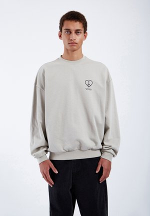 Light beige sweatshirt with ribbed cuffs and hem. Front features a small dark graphic in the shape of a heart and text underneath.