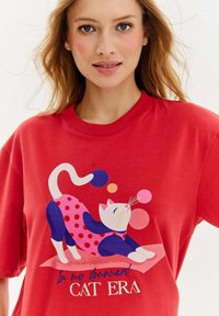 Czerwona koszulka z zabawnym nadrukiem kota w różowym ubranku w kropki. Zawiera tekst: "W mojej opadającej EPOCE KOTA." Krótkie rękawy, okrągły dekolt.
