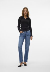 Crni longsleeve top s ovratnikom, uparen s plavim trapericama ravnog kroja srednje plave boje. Model nosi crne cipele s oštrim vrhom, a pozadina je bijela.