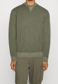 Mann in olivgrüner Langarm-Sweatshirt und Cargo-Hose, der mit entspannten Armen an den Seiten vor einem weißen Hintergrund steht.