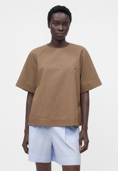 Γυναίκα που φοράει χαλαρό καφέ υφασμάτινο κοντομάνικο τοπ σε συνδυασμό με ανοιχτό μπλε υφασμάτινη σορτς μέχρι το γόνατο, στέκεται μπροστά σε ένα απλό φόντο.