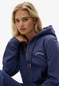 Granatowa bluza z kapturem na zamek, wykonana z miękkiej mieszanki bawełny, z kieszenią kangurką i białym haftowanym logo "SUPERDRY" na piersi.