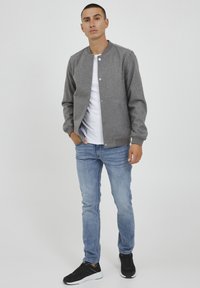 Grå bomberjakke med trykknapper, ribbestrikkede mansjetter og rund hals over en hvit t-skjorte, kombinert med lyseblå jeans og svarte sneakers.