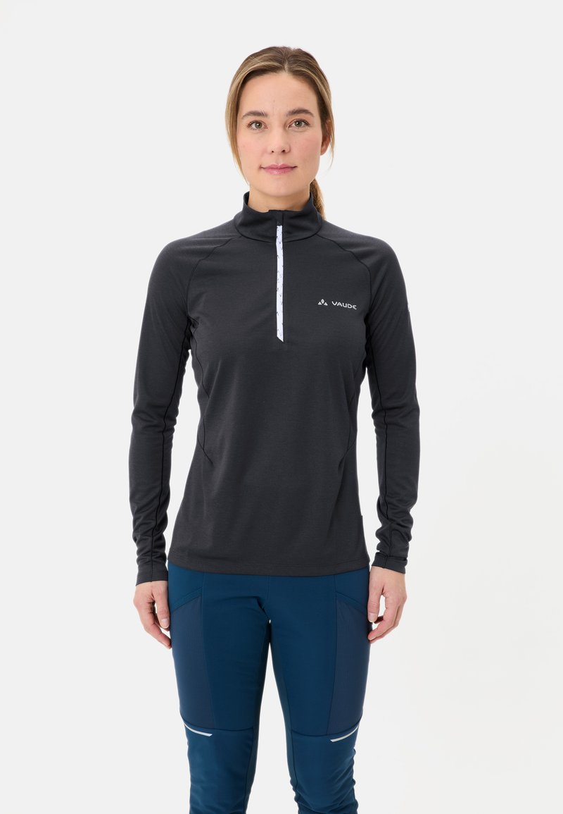 Frau in einem schwarzen langärmeligen, mit Reißverschluss versehenen Sportoberteil und blauen, engen Hosen, die vor einem schlichten weißen Hintergrund steht.