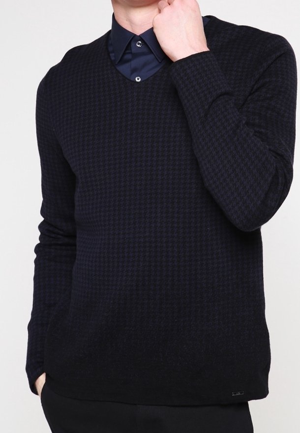 Homme portant un pull à manches longues texturé bleu marine foncé par-dessus une chemise bleu marine boutonnée, une main près du cou et l'autre dans la poche.