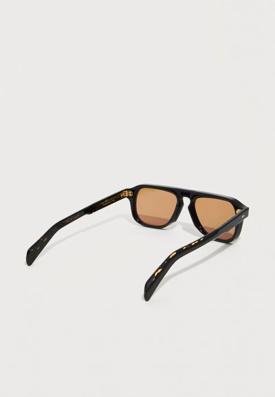 Occhiali da sole rettangolari neri con lenti marroni sfumate e aste decorate, poggiati aperti su una superficie bianca.