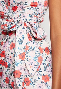 Robe portefeuille fleurie en tissu doux avec un fond rose orné de fleurs rouges et bleues, détail à nouer à la taille, et une texture lisse et légère.