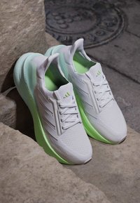 Paire de chaussures de sport blanches avec semelles et semelles intérieures vert clair placées sur des marches en pierre près d'une plaque d'égout.