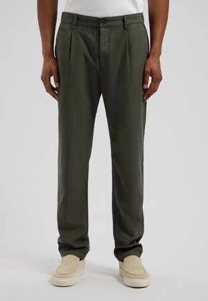 Osoba ubrana w ciemnozielone plisowane spodnie i beżowe buty wsuwane, stojąca na tle jasnoszarego, jednolitego tła.