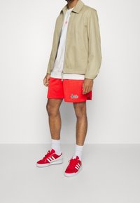 Beige vetoketjullinen takki, valkoinen T-paita, punaiset verkkomaiset shortsit logolla ja punaiset tennarit valkoisilla raidoilla ja nauhoilla. Valkoiset nilkkasukat.