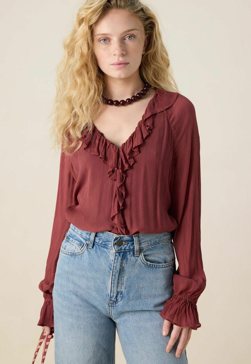 Blouse maroon à volants avec un décolleté en V et des manches longues, associée avec un jean en denim bleu clair taille haute. Texture de tissu lisse, coupe décontractée.
