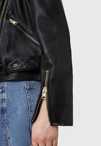 Giacca da biker in pelle nera con tasche e polsini con zip, dettagli in metallo dorato e finitura testurizzata. Primo piano sul dettaglio della manica.