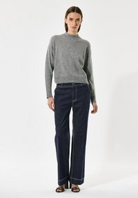 Sieviete, ģērbusies pelēkā adītā džemperī, tumši zilās platās biksēs un melnās atvērtās apavās ar papēdi, stāv uz vienkārša baltā fona.