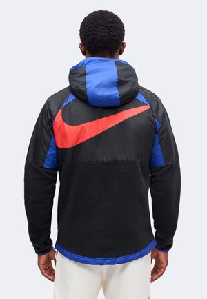 Homme portant une veste noire et bleue avec le logo rouge Nike dans le dos, debout dos à la caméra devant un fond uni.