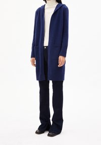Mörkblå huvtröja i mjukt material med öppen framsida, två framfickor och ribbade ärmar. Matchas med mörka utsvängda jeans och svarta skor.