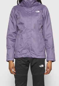 Personne portant une veste imperméable violette et un pantalon noir, tous deux arborant le logo The North Face, debout devant un fond uni.