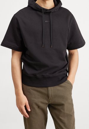 Mand iført sort kortærmet hættetrøje med "BOSS"-logo og brune ternede bukser, stående med den ene hånd i lommen og den anden afslappet.