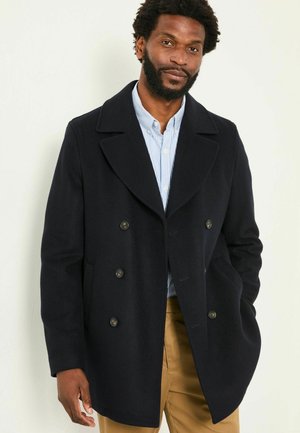 Navy peacoat with double-breasted buttons, wide lapels, and side pockets, made from a soft, textured fabric. Worn over a light blue striped shirt.