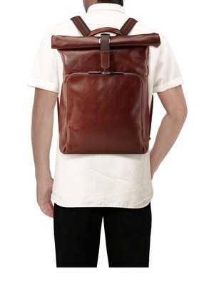 Man draagt een wit overhemd en zwarte broek, en heeft een grote bruine leren roll-top rugzak met een voorvak met rits.