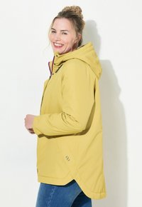 Gul vattentät jacka med huva, med en rundad nederkant, sidofickor och kontrasterande dragkedjor. Tillverkad av ett mjukt, lätt material.