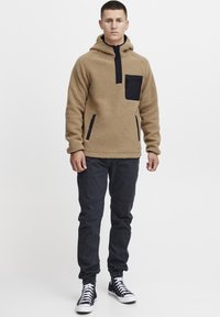 Tan fleece hættetrøje med en sort lynlåslomme på brystet, sidelommer og sorte detaljer. Kombineret med mørke bukser og sorte high-top sneakers.