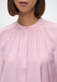 Lyserosa bluse med høy krage og rynkede detaljer ved utrigningen, med knappelukking og en lett rynket tekstur.