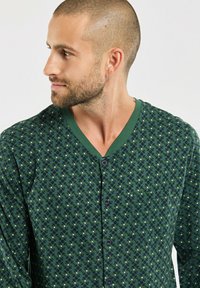 Chemise à boutons verte à motifs présentant un col en V, des manches courtes et une poche, ornée de petits motifs circulaires bleus et blancs sur le tissu.