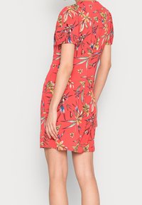 Femme portant une robe florale rouge avec des manches ballons courtes, vue de dos contre un fond gris clair uni.