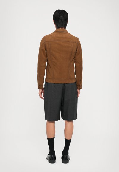 Brązowa zamszowa kurtka o dopasowanym kroju, z dwoma bocznymi kieszeniami i subtelnymi detalami szycia, zestawiona z szarymi szortami i czarnymi butami.