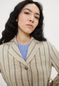 Beige- und blau gestreifter Blazer mit Knopfverschluss, zwei Taschen und Reverskragen. Getragen über einem hellblauen Strickpullover.