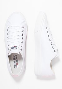 Paar weiße Low-Top-Canvas-Sneaker mit weißen Schnürsenkeln, Metallösen und "Tommy Jeans"-Markenaufdruck auf der Innensohle und der Zunge.