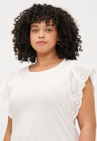 A person with curly hair wears a white shirt featuring detailed eyelet ruffled sleeves, facing the camera with a neutral expression.
