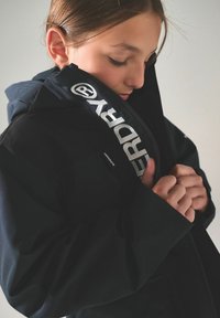 Svart vattentät jacka med dragkedja, som har en hög krage och logodetaljer. Materialet verkar slätt med en subtil lyster.