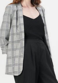 Šedý a černý kostkovaný blazer se uvolněným střihem, v kombinaci s černým topem s výstřihem do V a černými kalhotami s vysokým pasem. Hladké textury, žádné viditelné kování.