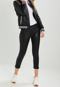 En ung kvinna står självsäkert, iklädd en läderbombarjacka, åtsittande svarta byxor och vita sneakers, framför en neutral bakgrund.