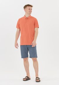 Korallfärgad polo med korta ärmar och tre knappar, tillsammans med blå shorts med rullad fåll och bruna sandaler med spänndetalj.