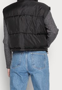 Black puffer vest with a shiny texture, featuring horizontal seams. Worn over a gray ribbed sweater and paired with light blue denim jeans.